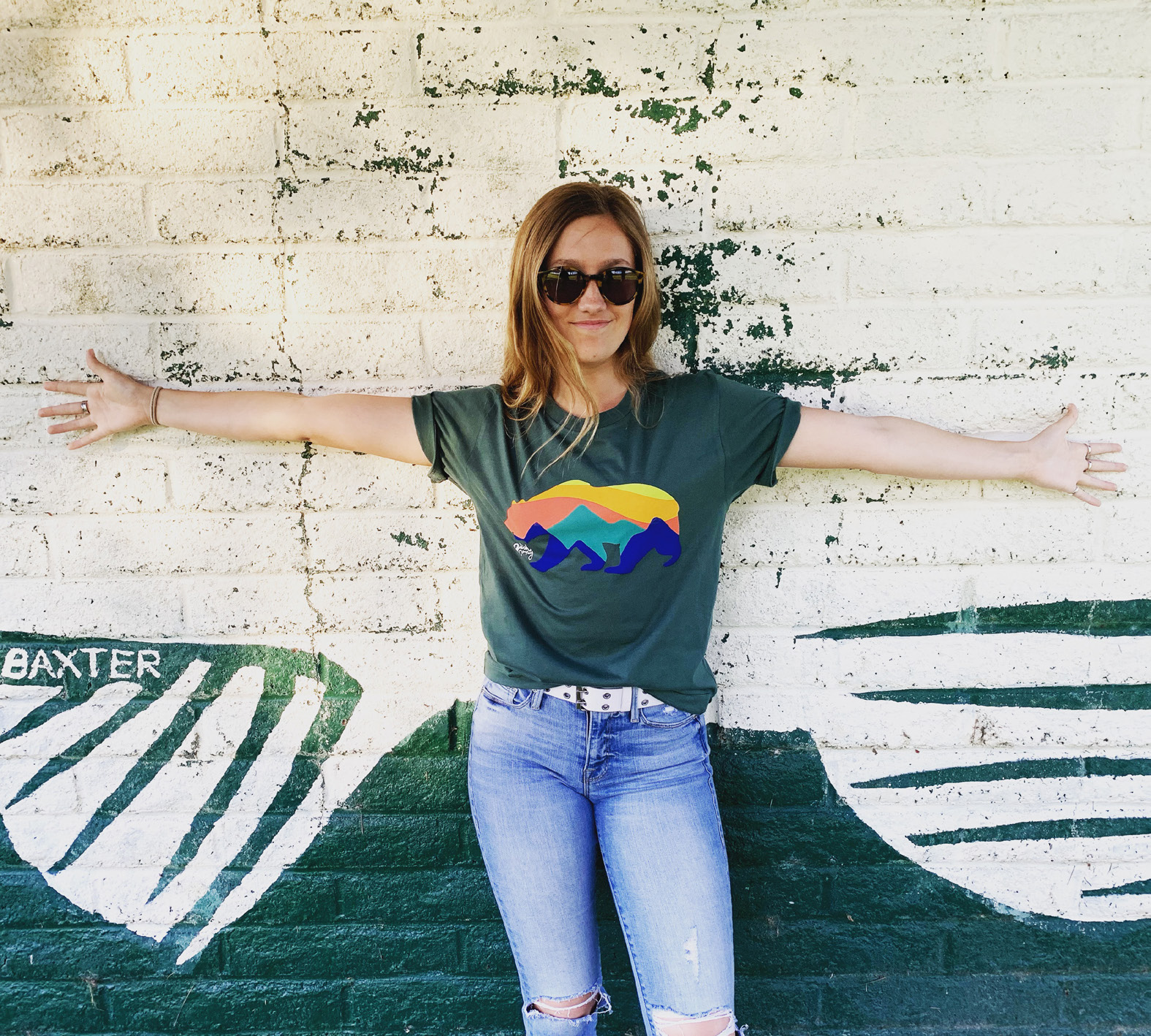 Lifestyle photograph of a woman wearing the green Sunrise Bear tee against a painted wall