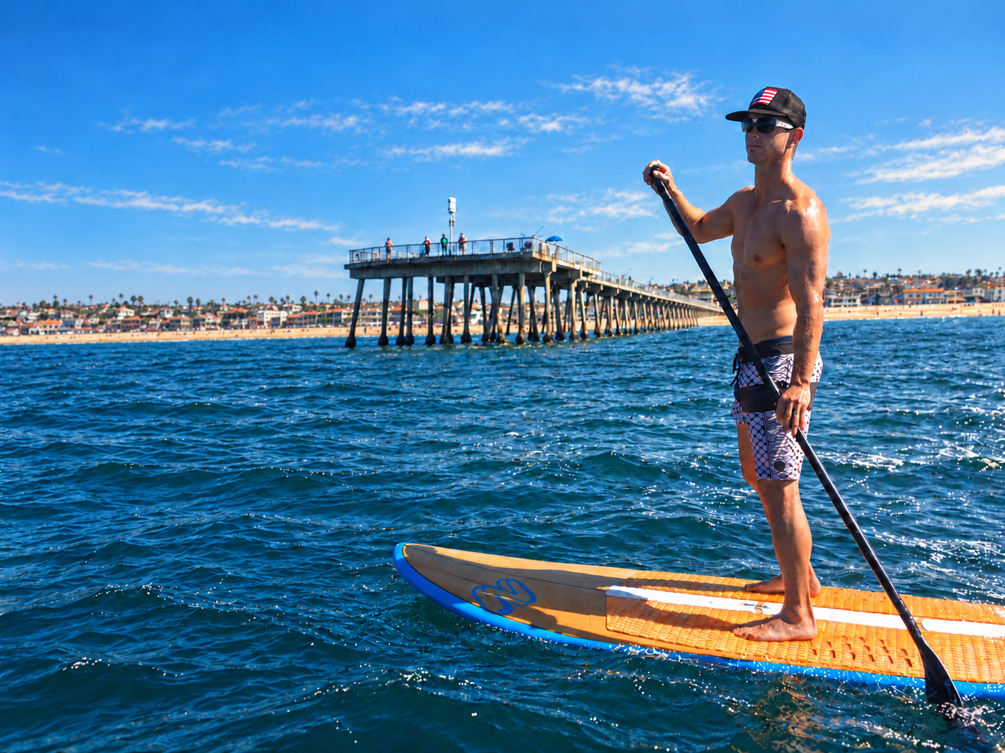 Man wearing a black American flag patch hat while paddleboarding beside a California pier